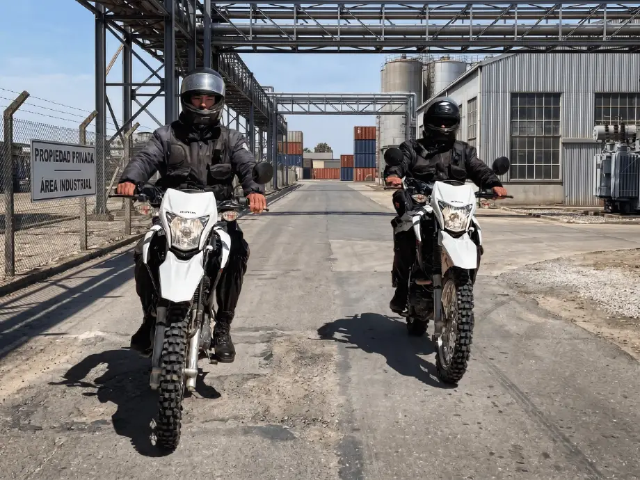 Dos vigiladores de GSP patrullando en moto el perímetro de una planta industrial, junto al cartel de Propiedad Privada · Área Industrial