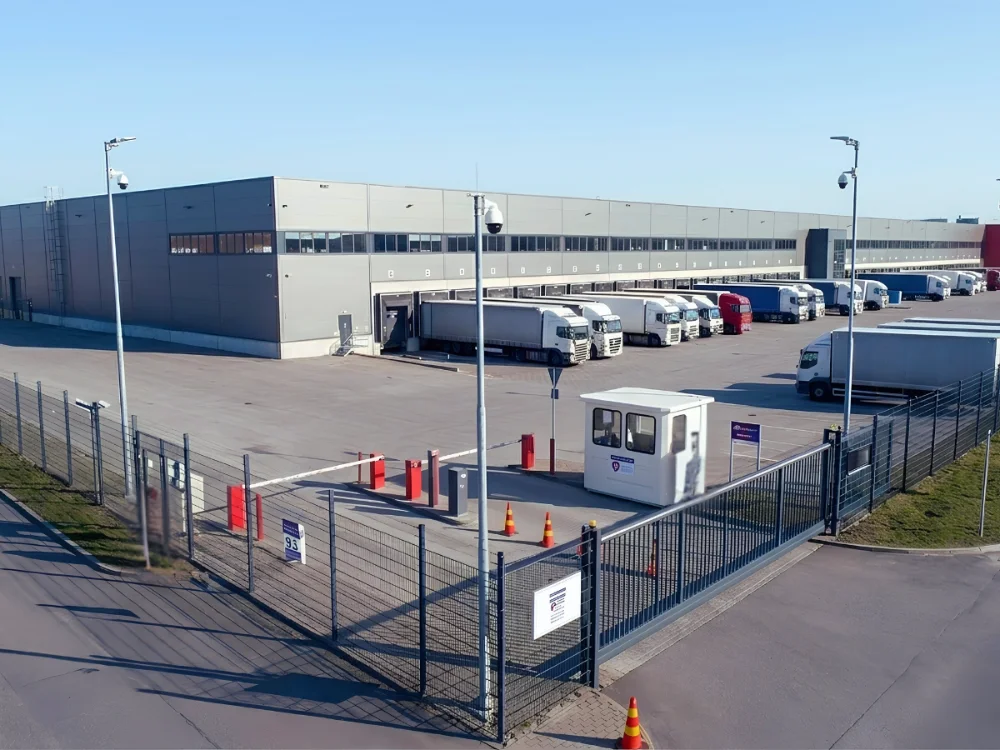 Planta logística industrial con guardia en garita, barrera de control de accesos vehicular y cámaras de seguridad